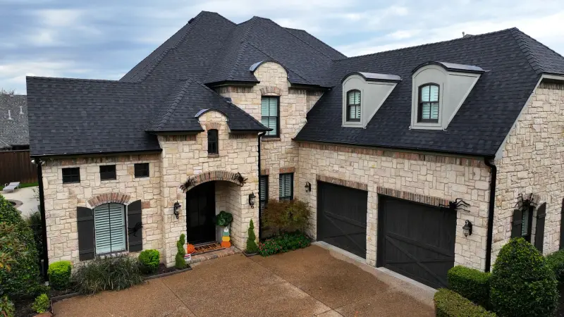 Drone view of a finished dark shingle roof on a two-story stone home with three dormers and black garage doors