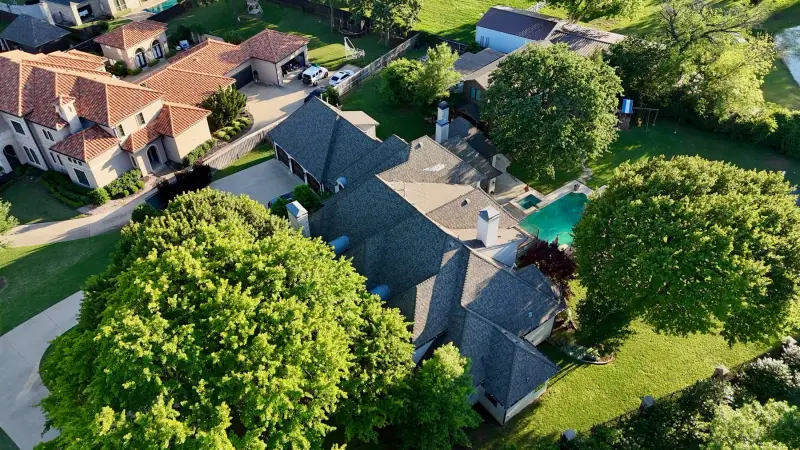 Drone view of a finished gray shingle roof on a stone and brick home surrounded by oak trees, with a pool in the backyard